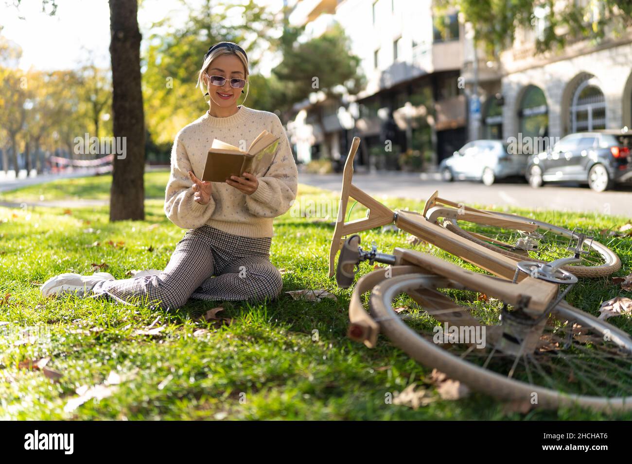 Full body of positive young female enjoying reading book while sitting on lawn near timber bicycle Stock Photo