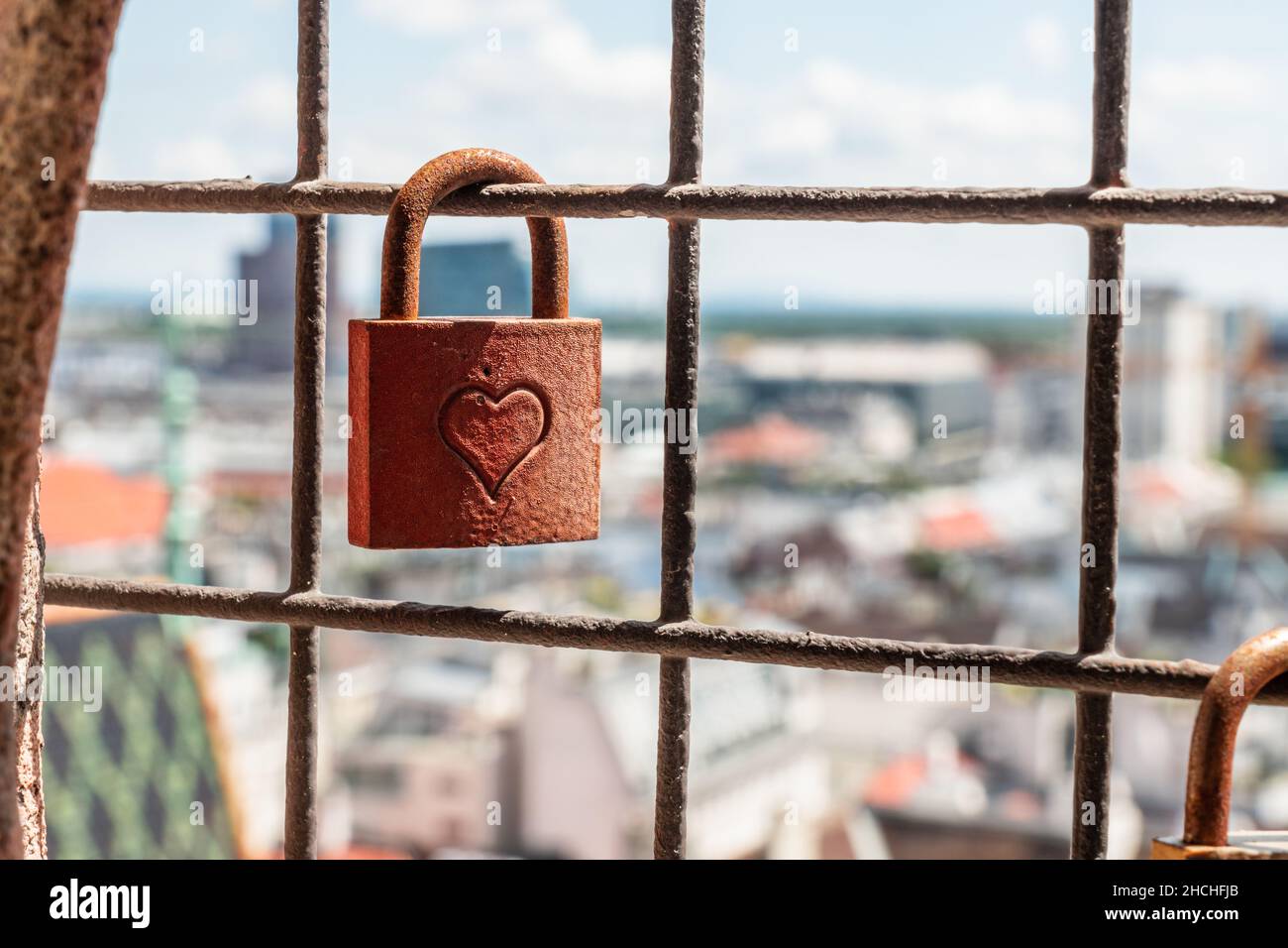 a rusted metal padlock hanging on a protective fence with a city view in a background Stock Photo