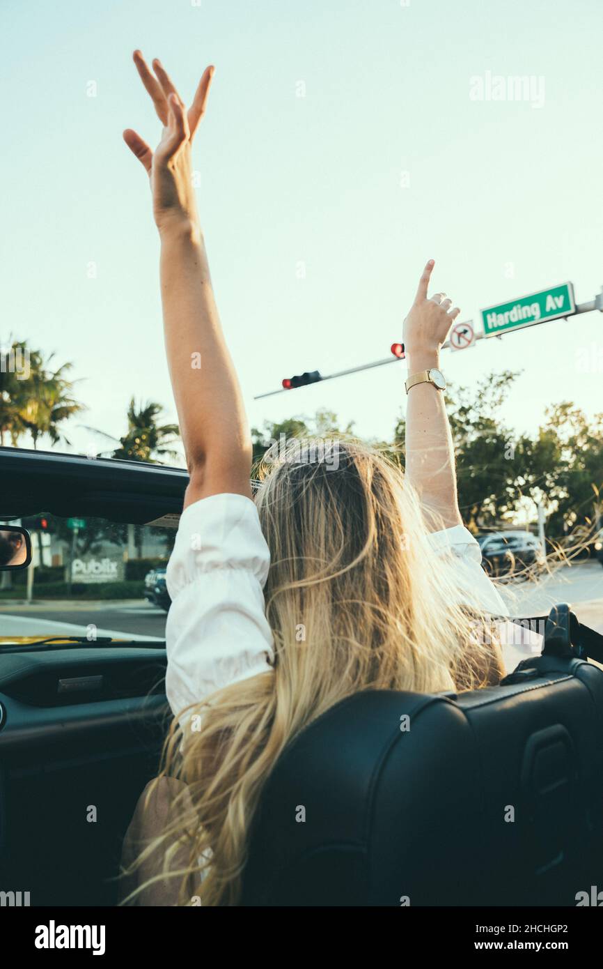 Vertical shot of a blonde girl having fun with her hands up in an open roof car Stock Photo