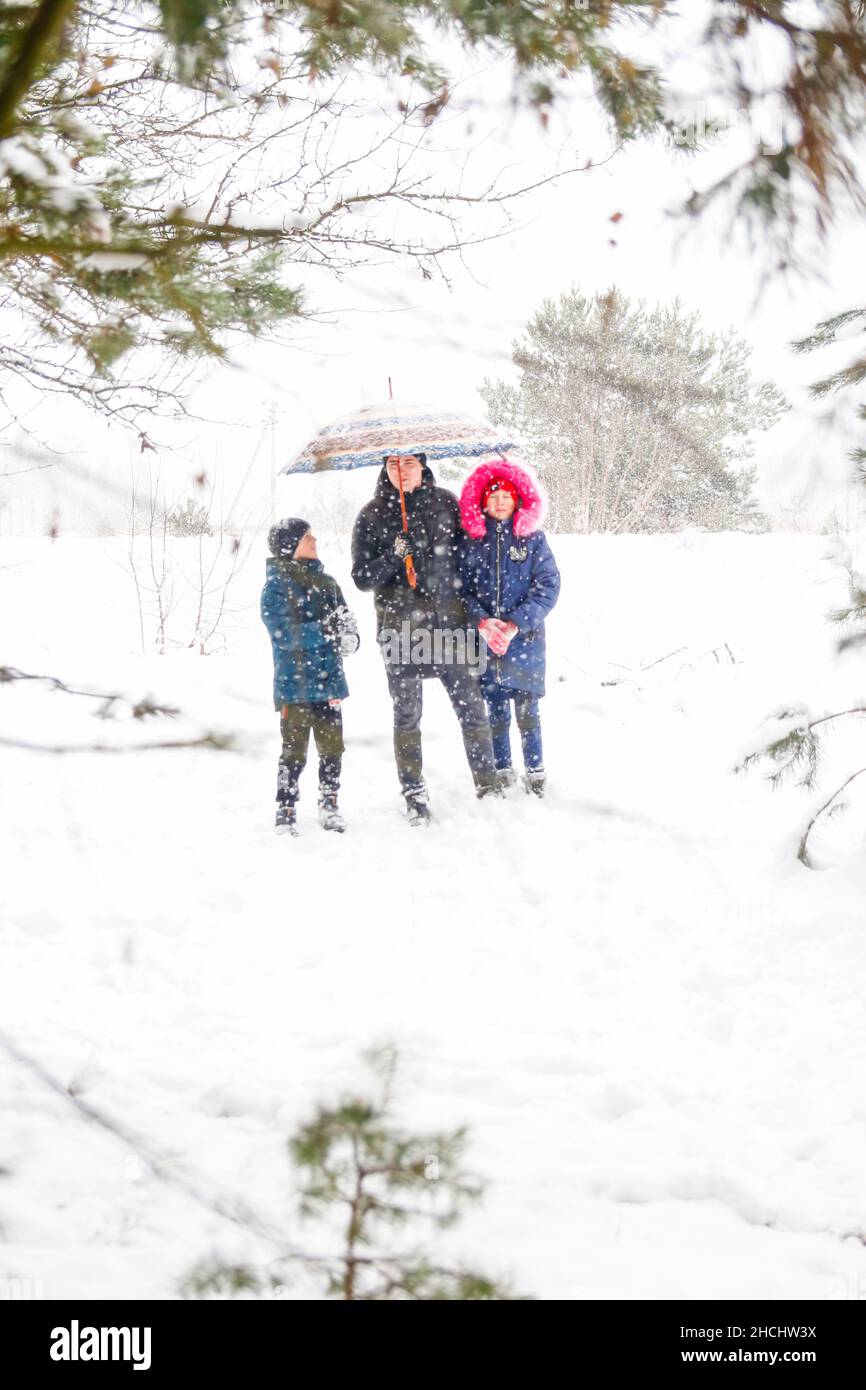 Young man in warm hat and child girl and boy on rural winter snowy background holding umbrella. Happy family, cold weather. Time together. Vacations. Stock Photo