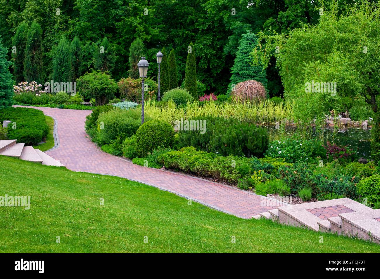stone steps descent to a path from paving slabs in park with iron retro lantern on slope with scenic plants, evergreen bushes and pine and foliage tre Stock Photo