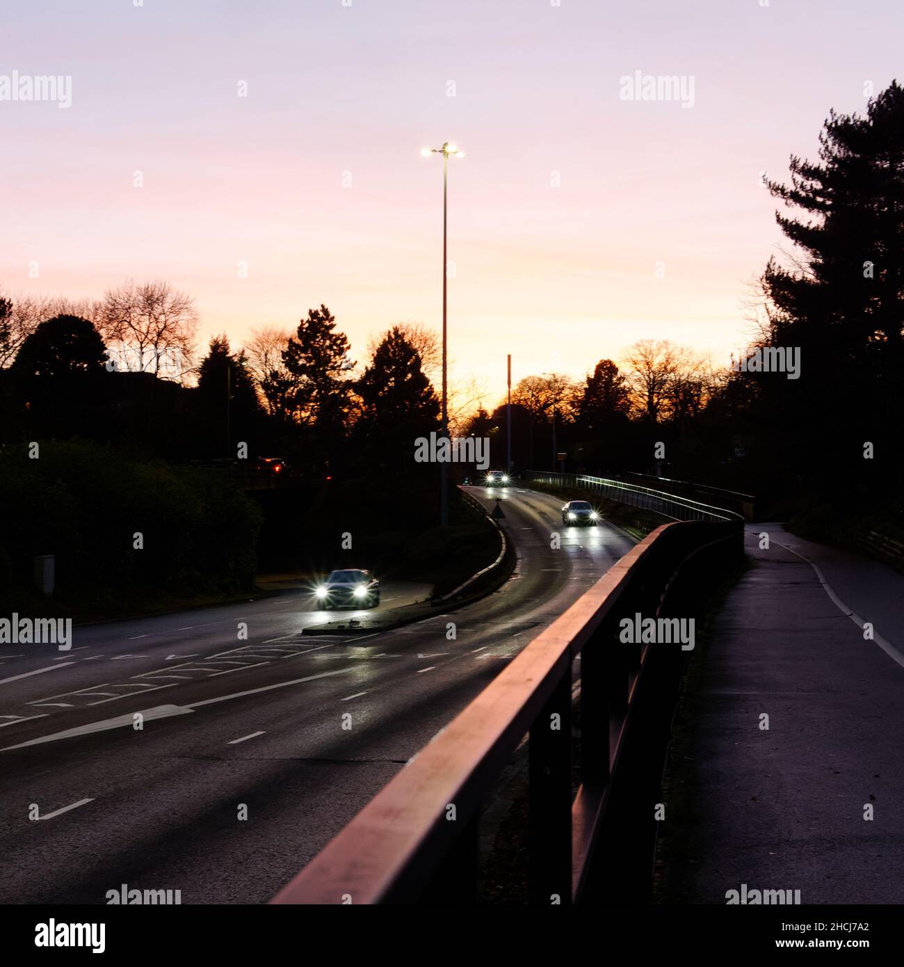 Cars driving on a slip road with lights on which form starbursts as the sun sets behind and reflects on the roadside barrier. Stock Photo