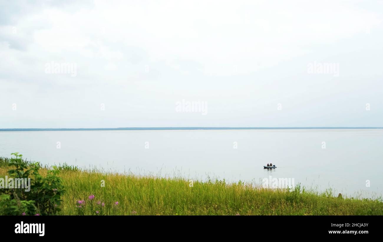 Green field and a calm lake with floating rubber boat. Fresh green grass and flowers on the shore of the big lake on cloudy sky background in the summ Stock Photo