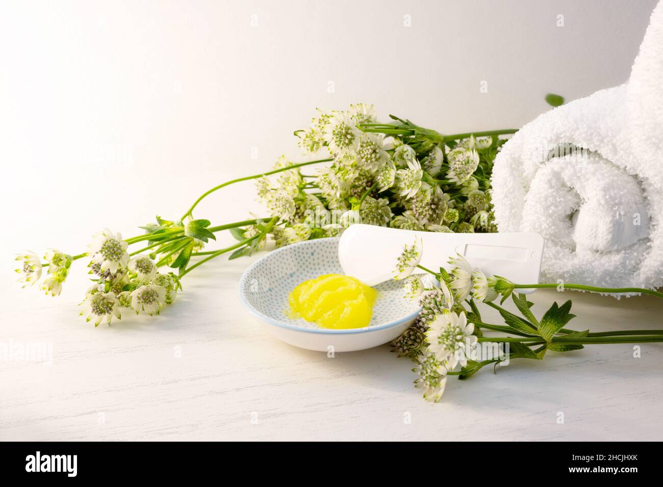 Cosmetic moisturizer or emollient and a spatula for pedicure and soft foot skin in a small bowl and some green flowers on a light wooden background la Stock Photo