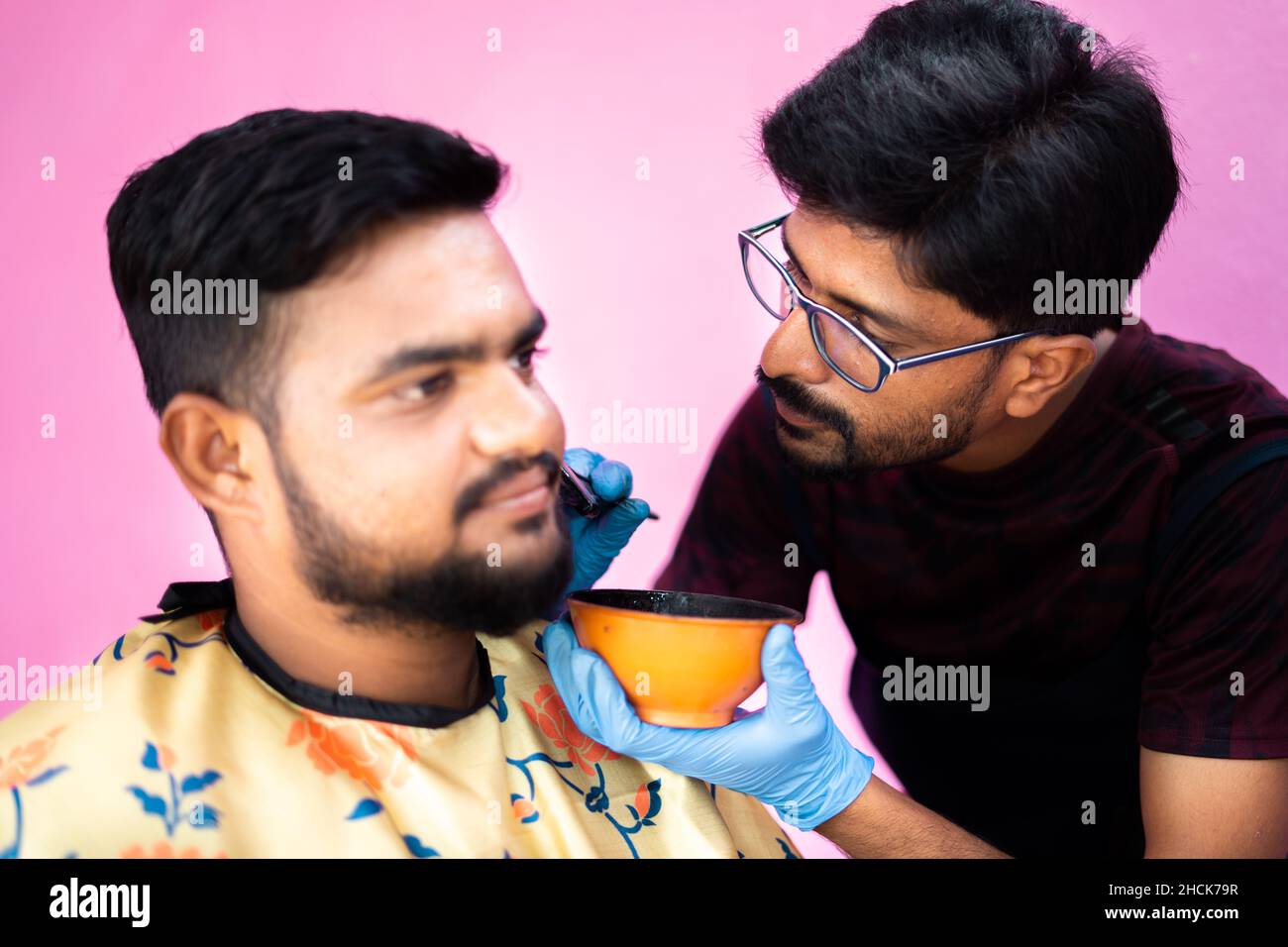 focus on barber, close up shot of concentrated hairdresser applying hair dye or black henna to young man to cover white hair beard at salon - concept Stock Photo