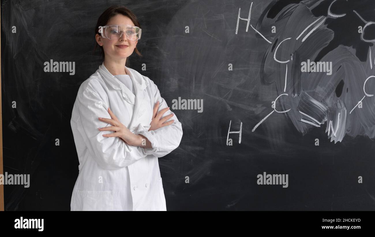 A young girl in a white coat and goggles is standing against the background of the blackboard with the chemical formula. Chemistry teacher or student Stock Photo