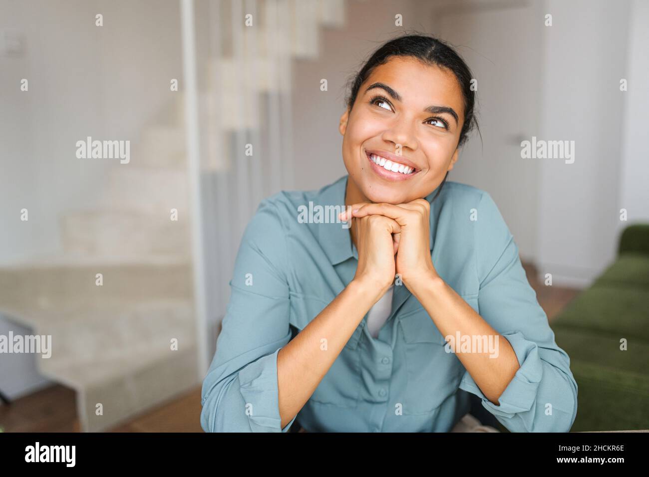 Headshot of young dreaming African American ethnic female with beautiful smile and looking away while sitting against blurred home or office interior background Stock Photo