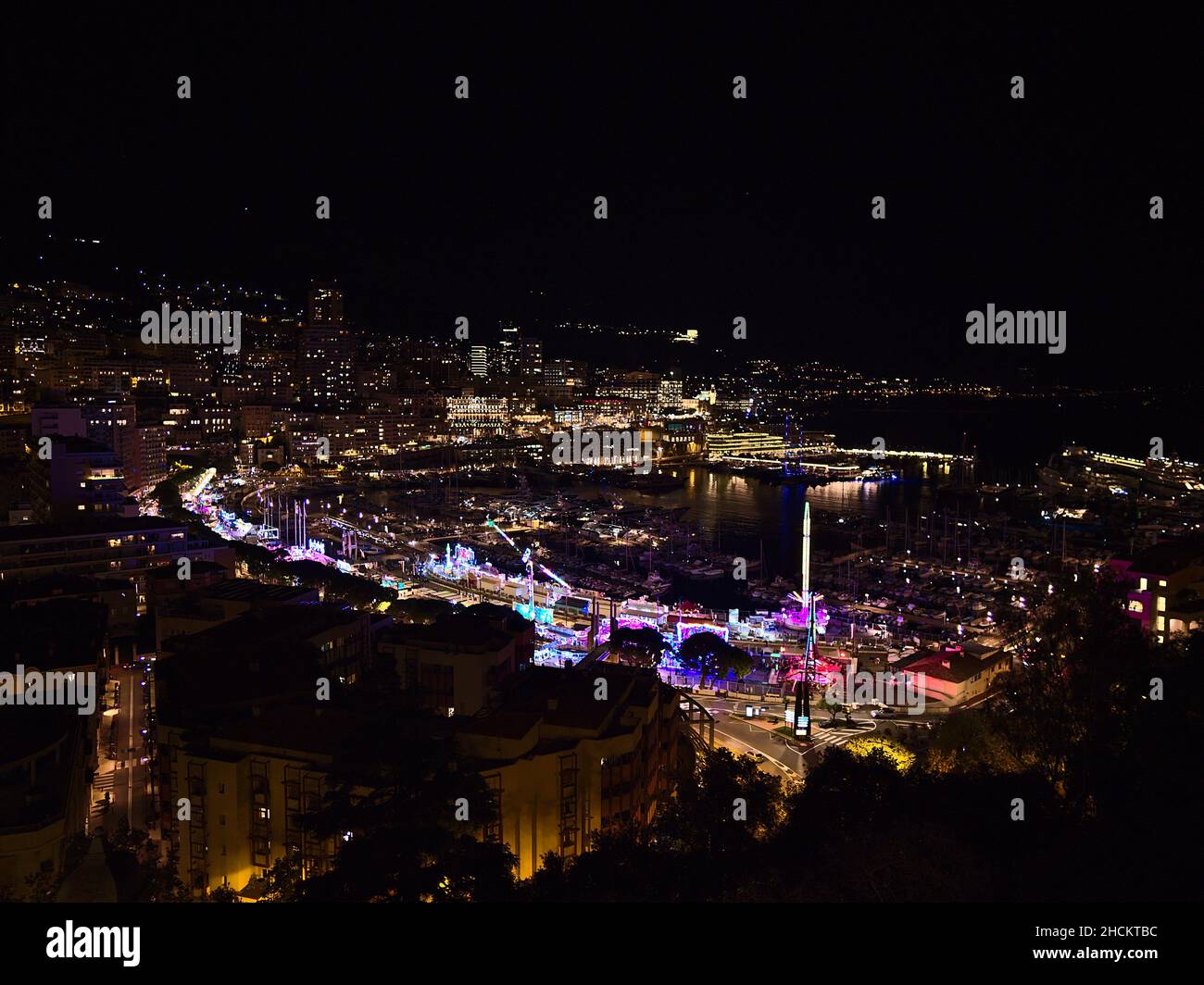 Beautiful panoramic view of the illuminated downtown of Monaco at night with colorful funfair and marina Port Hercule surrounded by buildings. Stock Photo