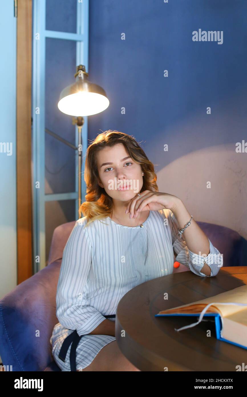 One young adult pretty female girl looks at camera, holds hand near face and sits in chair at table on background of lamp and blue walls in stylish Stock Photo