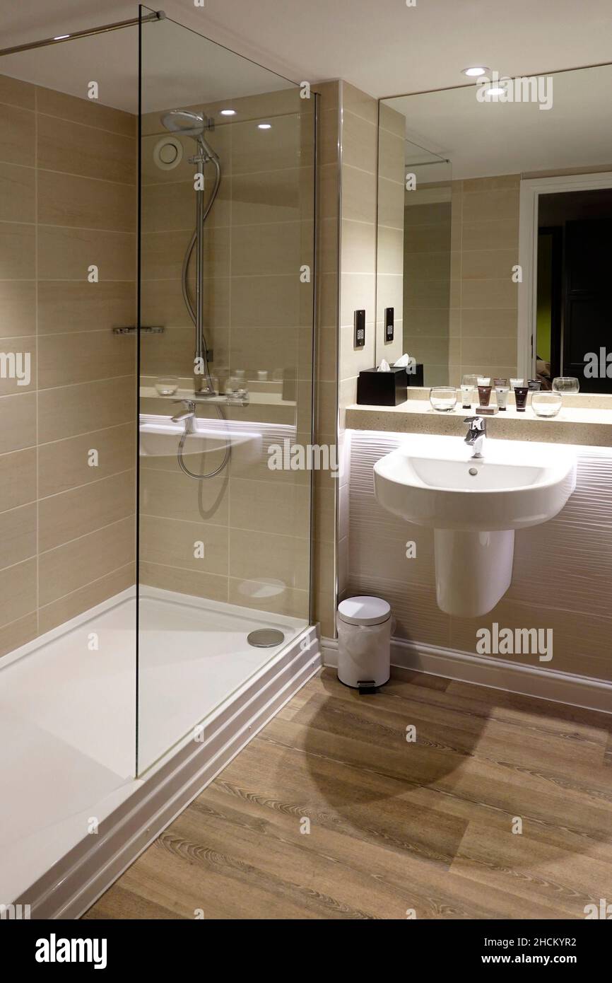 Modern walk in tiled shower & screen large safety grab rail in en suite hotel bathroom full height mirror over wall mounted wash hand basin England UK Stock Photo