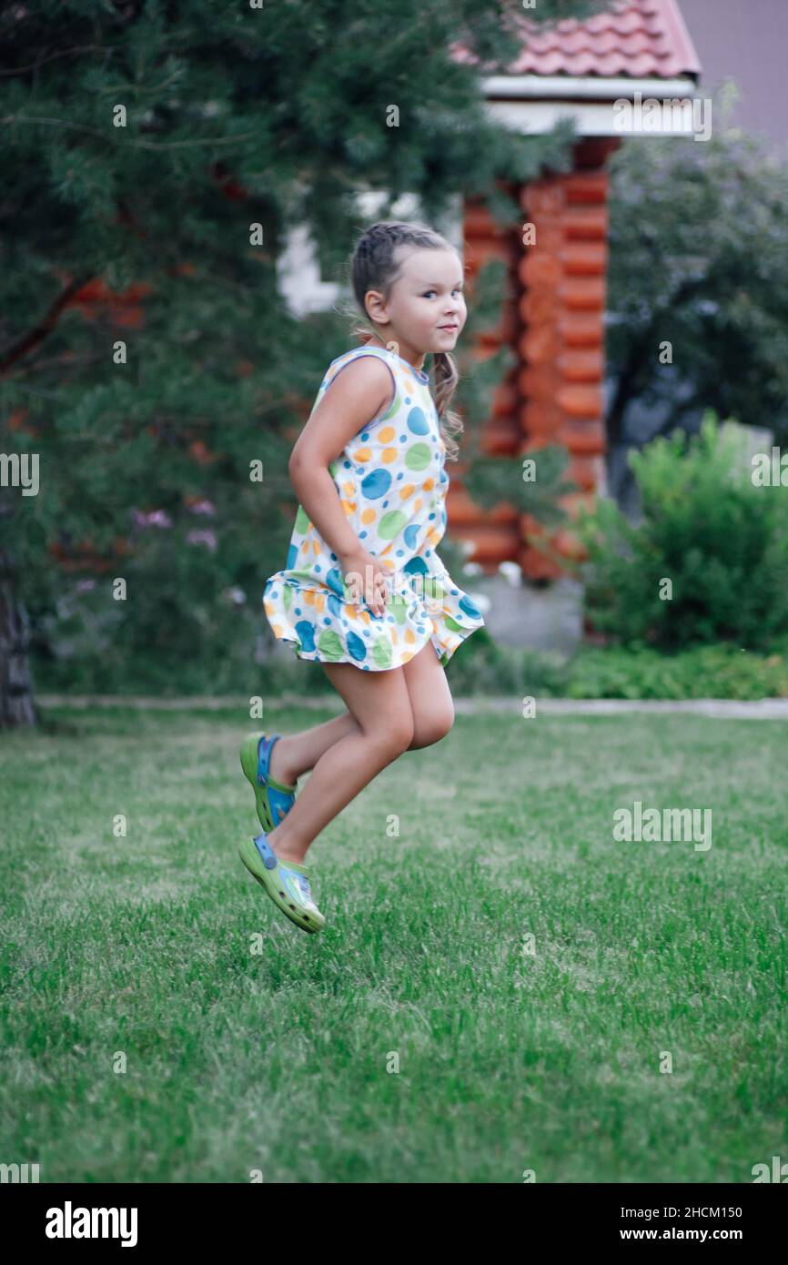 Pretty little girl in jump smiling, looking at camera having fun wearing light summer dress and slippers in park in daytime. Amazing background full Stock Photo