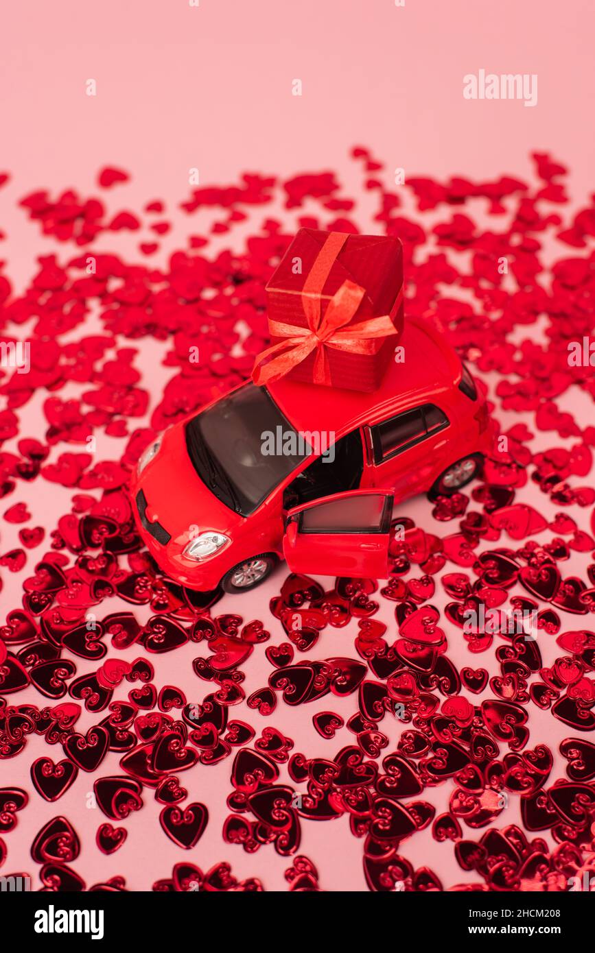 high angle view of toy car with wrapped present near shiny red confetti hearts on pink Stock Photo
