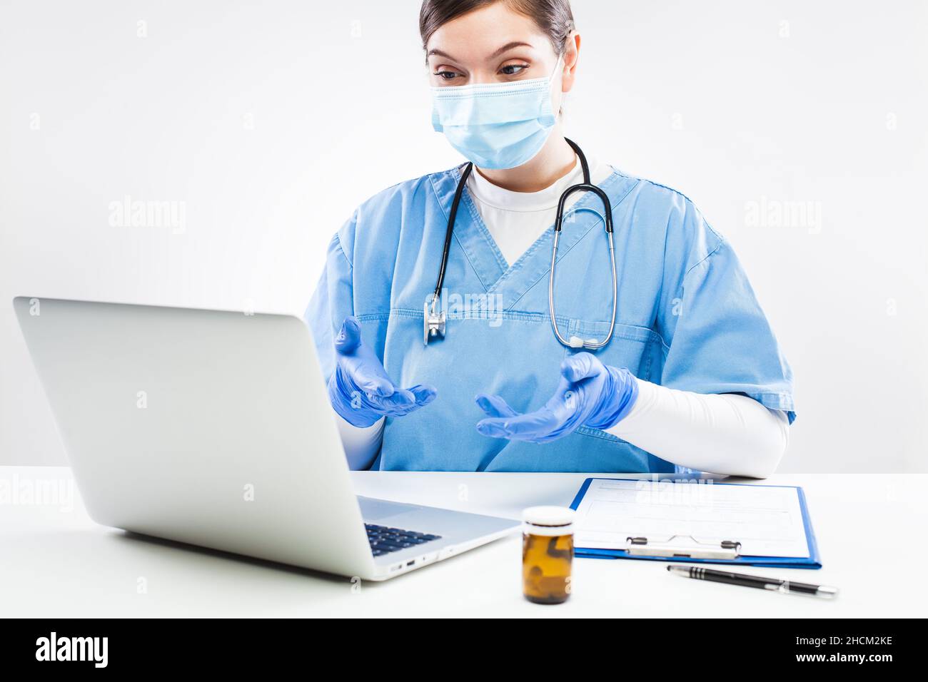 Young female caucasian NHS GP doctor sitting at desk in office,wearing blue uniform,protective face mask and gloves,virtual patient tele visit Stock Photo