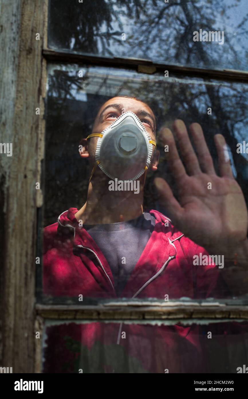 Young man wearing protective N95 face mask,looking through window from his room,hand on glass,depressed and anxious in COVID-19 lockdown quarantine Stock Photo