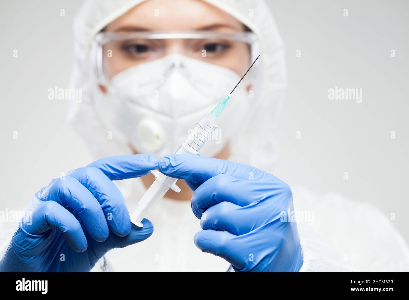 Female medical NHS worker wearing protective suit,goggles,face mask and gloves,holding syringe with needle containing vaccination booster dose Stock Photo