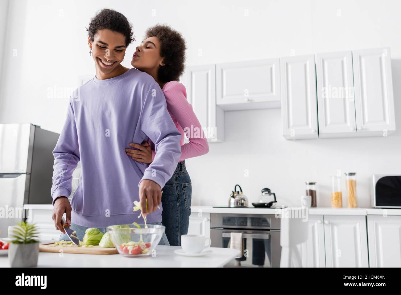 African american woman hugging and pouting lips near boyfriend cooking ...