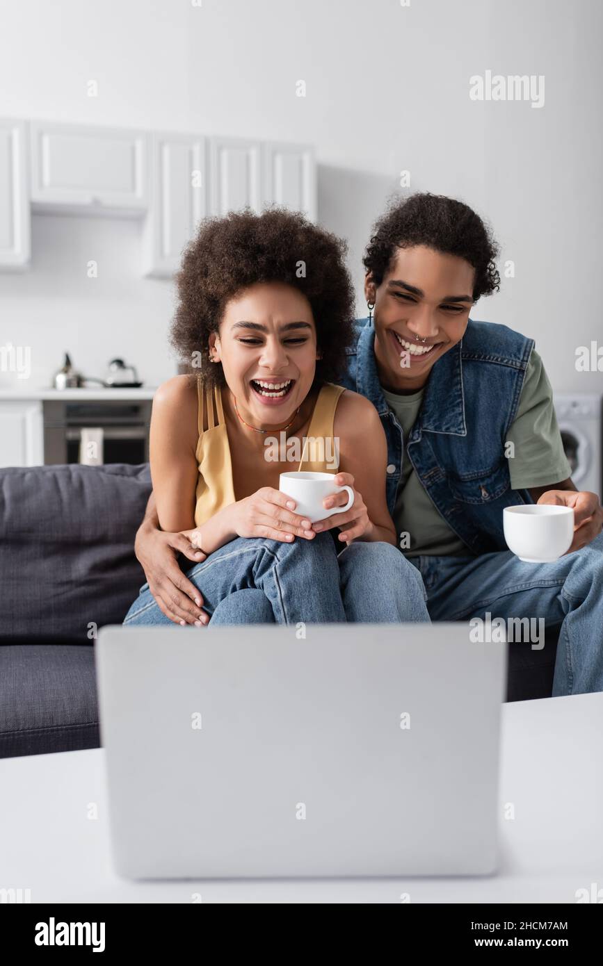 Cheerful african american couple with cups looking at blurred laptop at ...