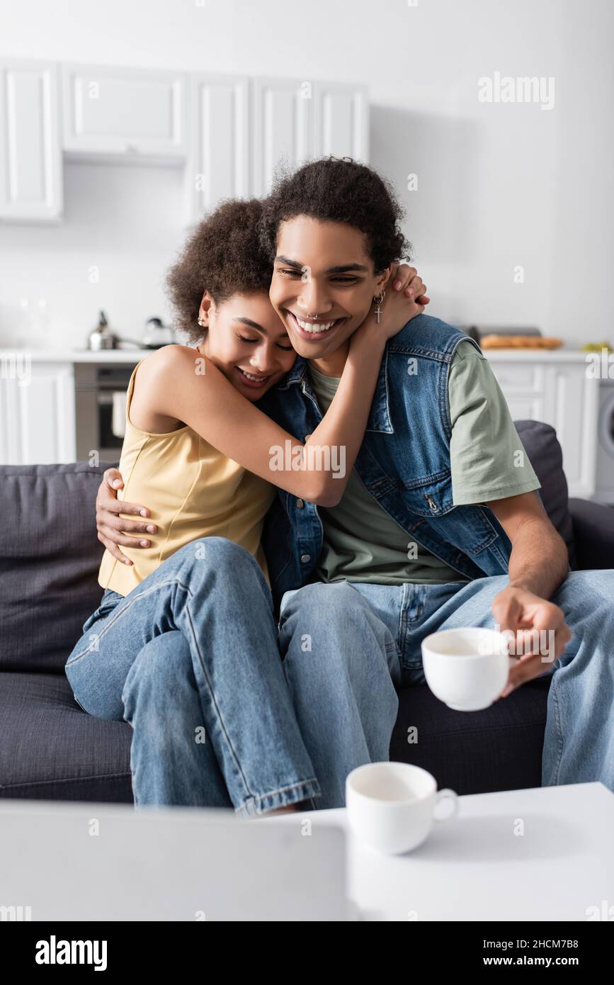 Cheerful african american woman hugging boyfriend with cup near laptop ...