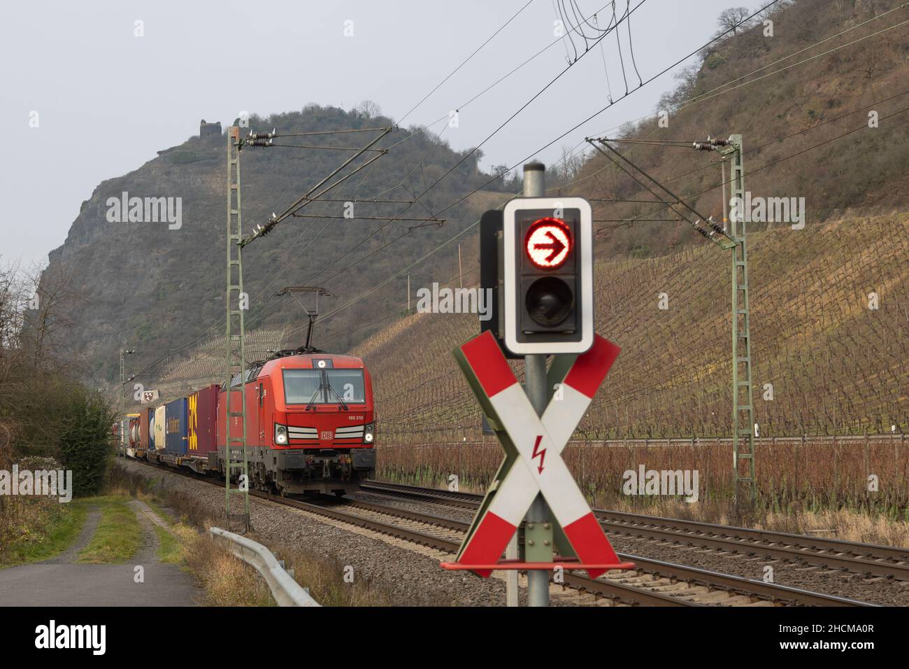 A freight train with a red electric locomotive and a railroad crossing signal with red lights in front Stock Photo