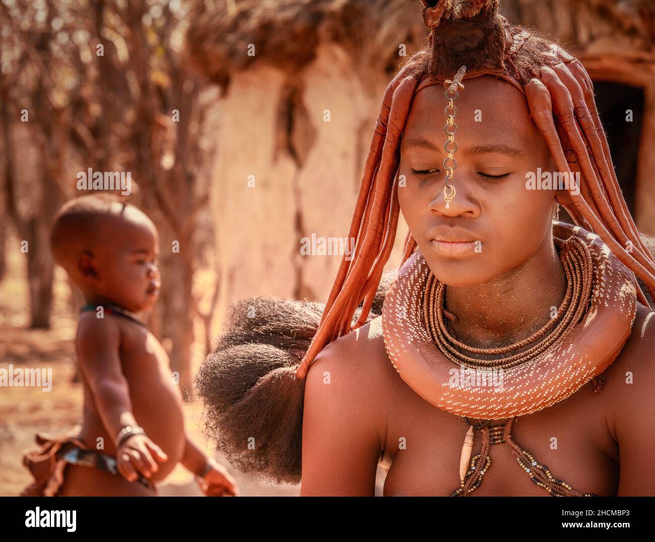 An African woman wearing traditional Himba tribal jewelry and hairstyle with her young child in the background in rural Namibia. Stock Photo