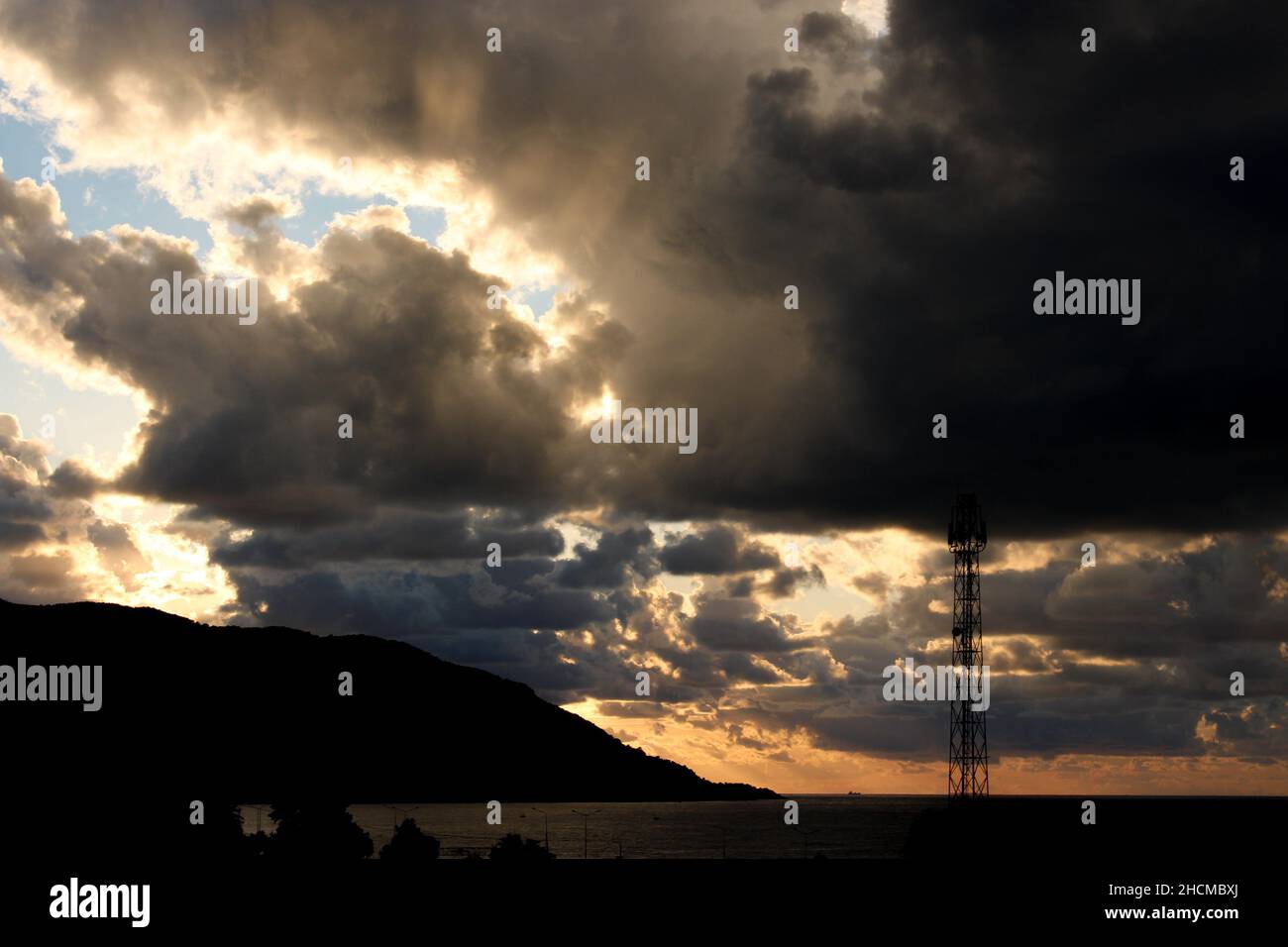 Dark storm clouds at sunset, dark storm clouds with sunrays, mountain and radio tower silhouette over the sea. Climate and storm concept Stock Photo