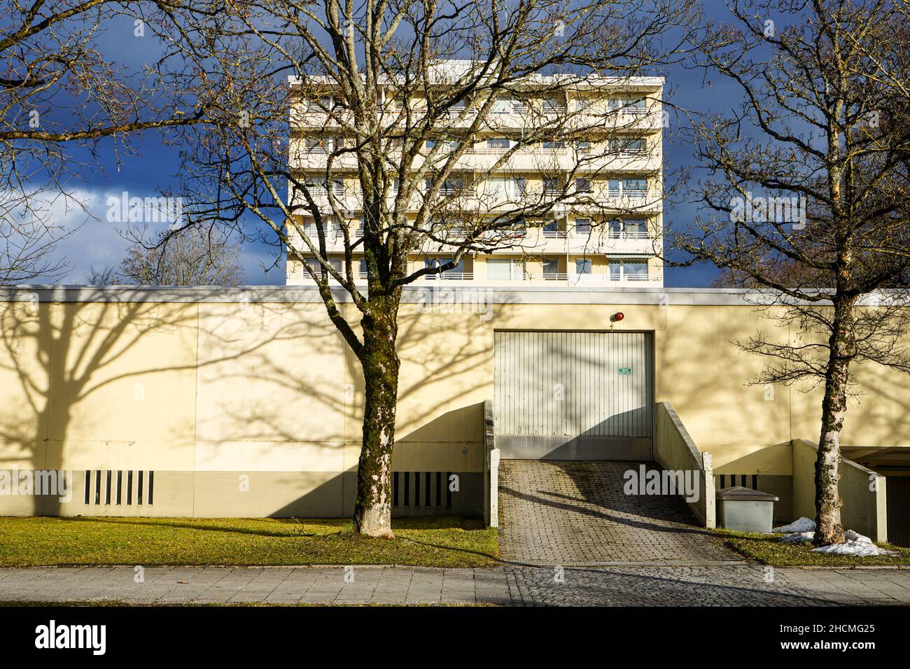 A flat elongated garage building with a gate. On the beige wall shadow reflections of trees. Behind it a tall apartment building complex. Stock Photo