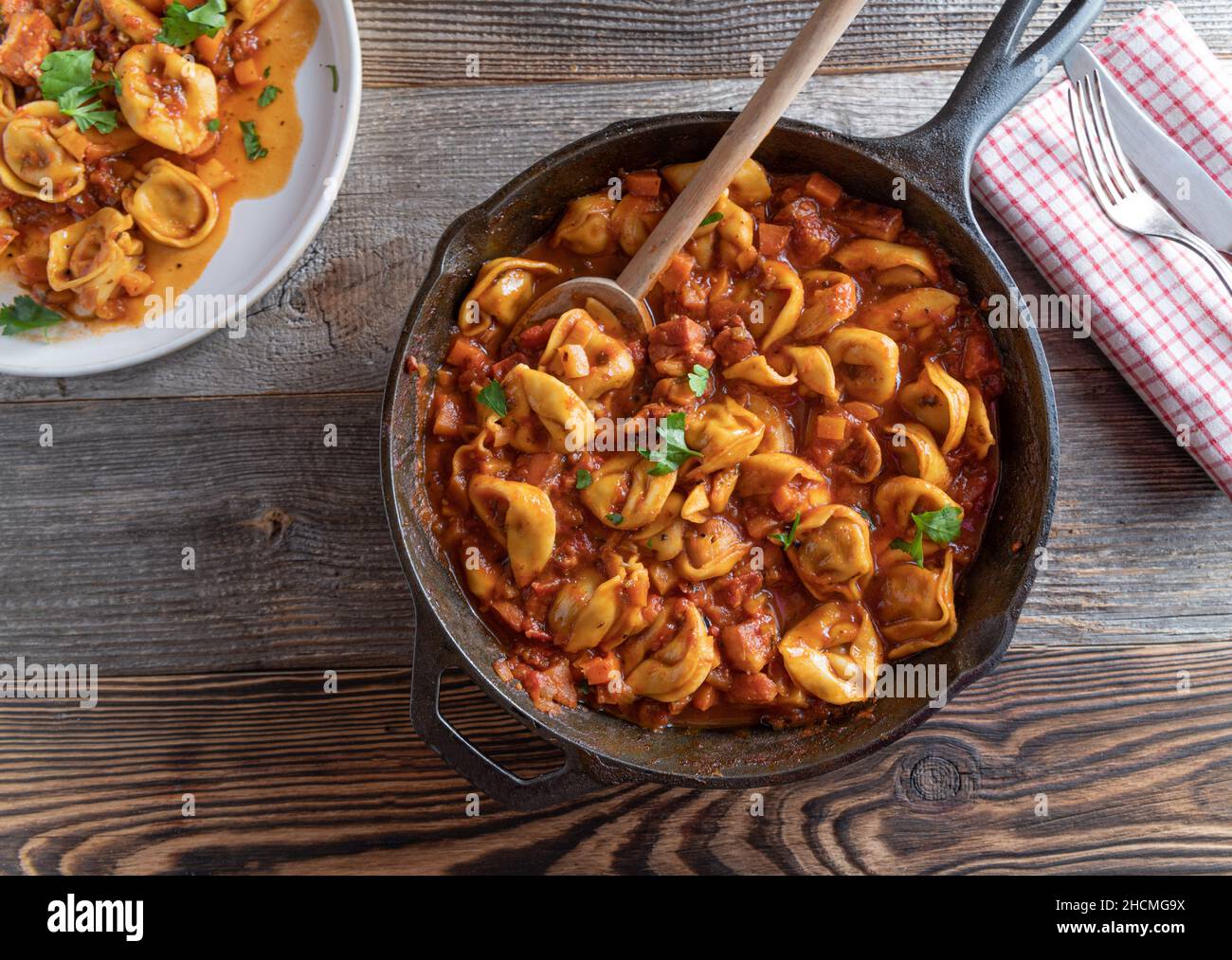 Italian pasta dish with tortellini in a delicious tomatoe, vegetable bacon sauce. Served in rustic pan isolated on wooden table background. Overhead Stock Photo