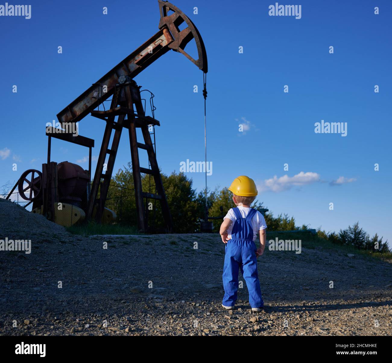 Back view of intelligent child studying business development at oil station with mining platform and drilling rig, boy in yellow helmet and blue uniform for kids looking at machine construction Stock Photo