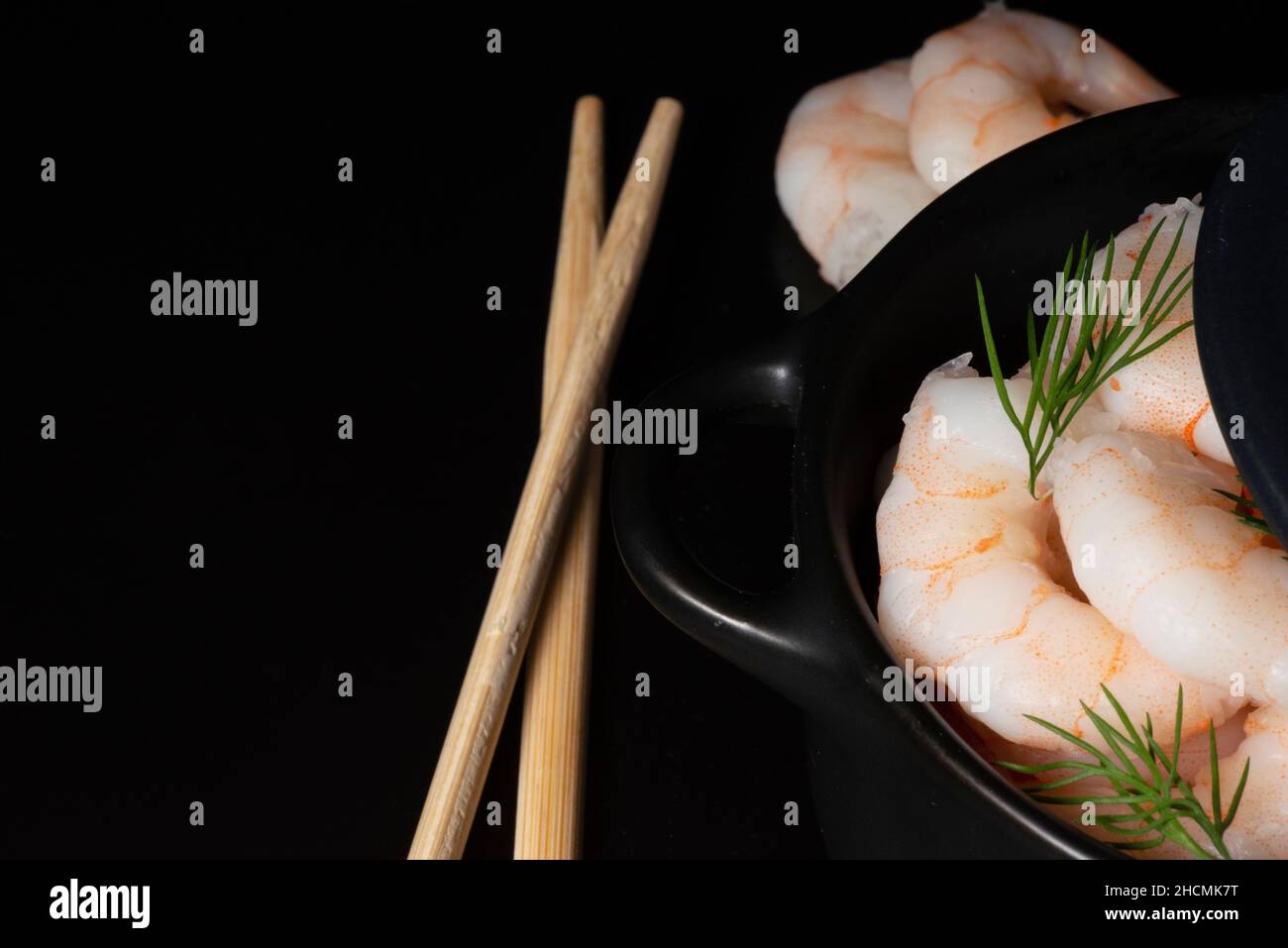 Close up side view detail of fresh cooked peeled prawns or shrimps in dark bowl and chop sticks on black background Stock Photo