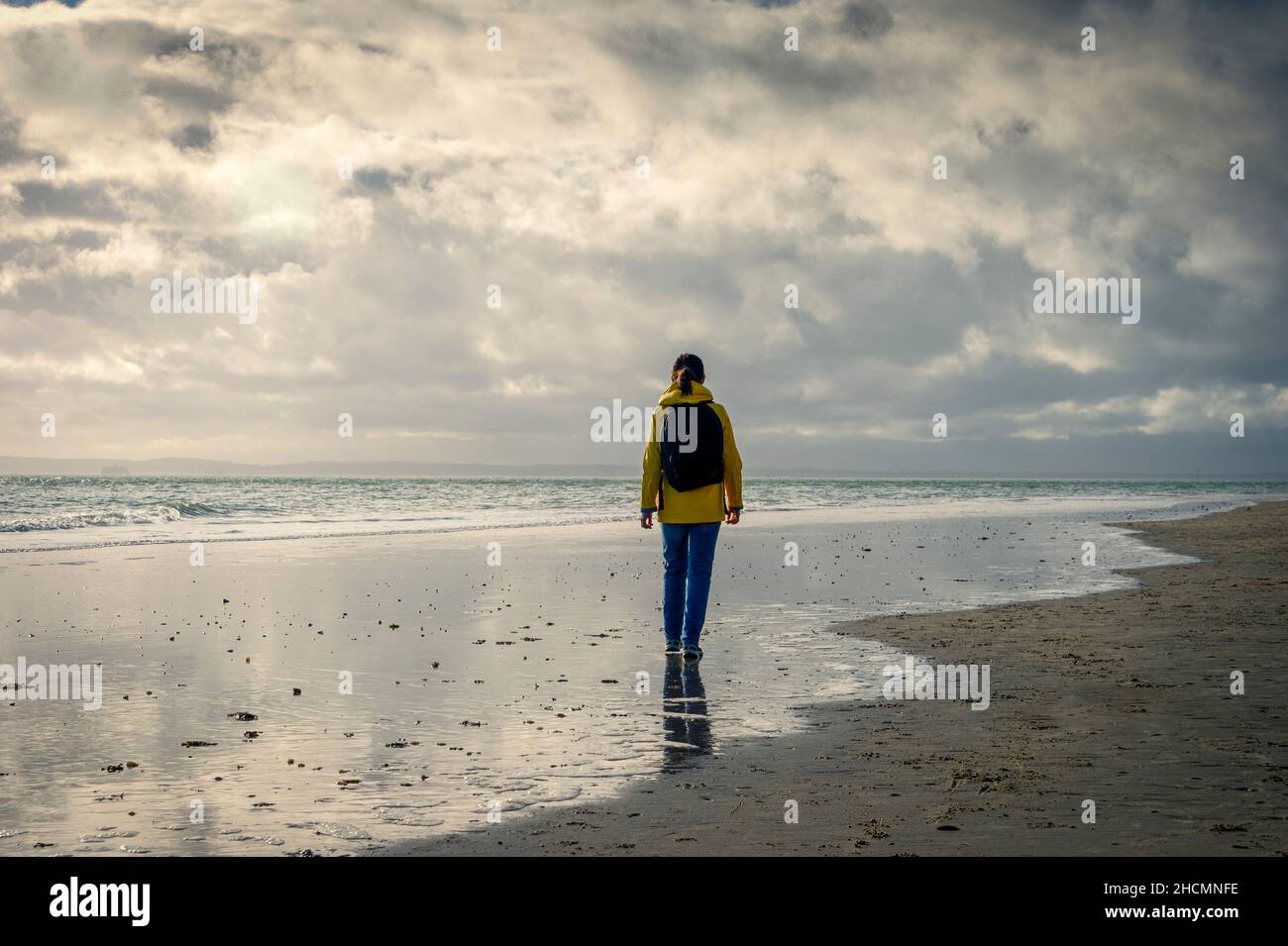 Rear view of a woman wearing a yellow jacket and backpack walking along the wet sand at the edge of the sea. Stock Photo