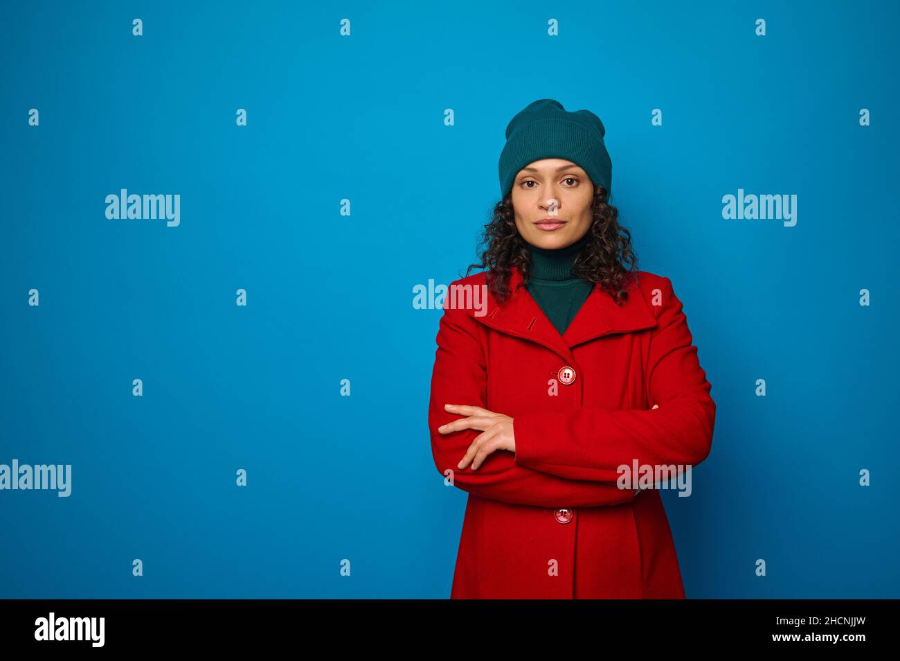Confident lifestyle portrait of a beautiful curly dark-haired pretty woman in red coat and green looking at camera posing with crossed arms against bl Stock Photo