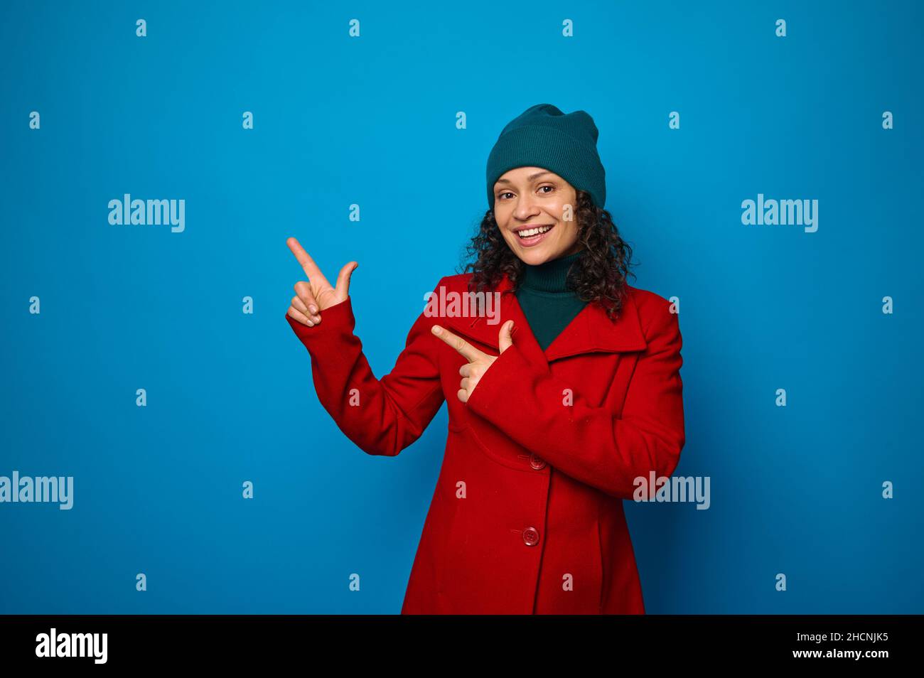 Charming smiling curly dark-haired woman in bright red coat and woolen green hat poses against blue background, looks at camera and points with finger Stock Photo