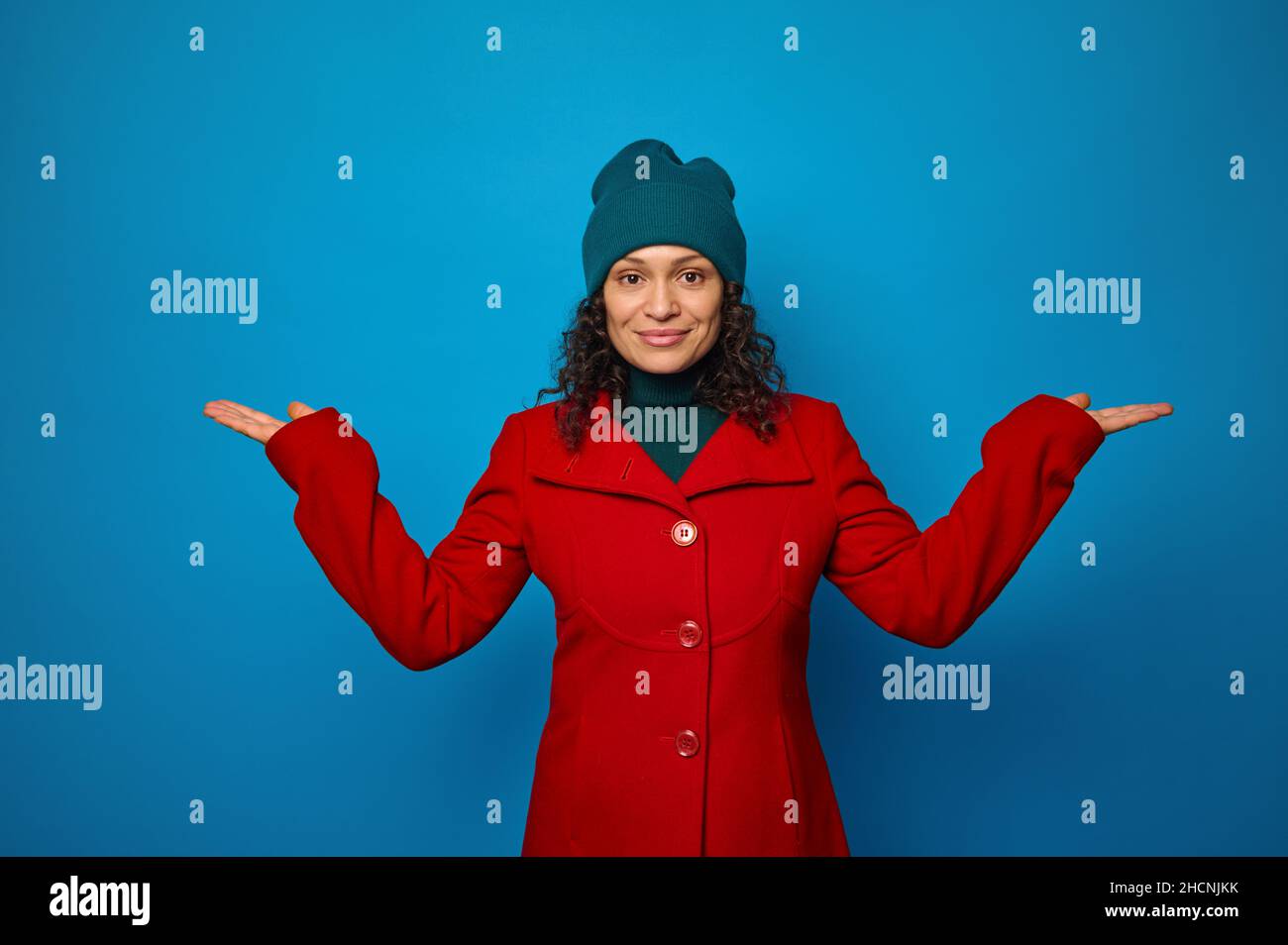 Conceptual portrait of a beautiful young curly haired brunette woman in red coat and green hat poses against blue background, looks at camera holding Stock Photo