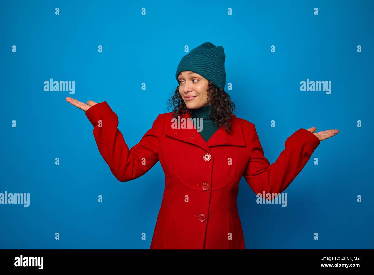 Conceptual portrait of a charming smiling curly dark-haired woman in bright red coat and woolen green hat poses against blue background, looks at an i Stock Photo