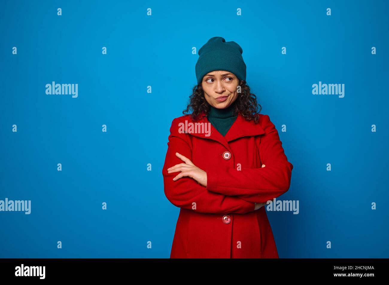 Portrait of a puzzled thoughtful pensive curly dark-haired pretty woman in red coat and green looking aside posing with crossed arms against blue wall Stock Photo
