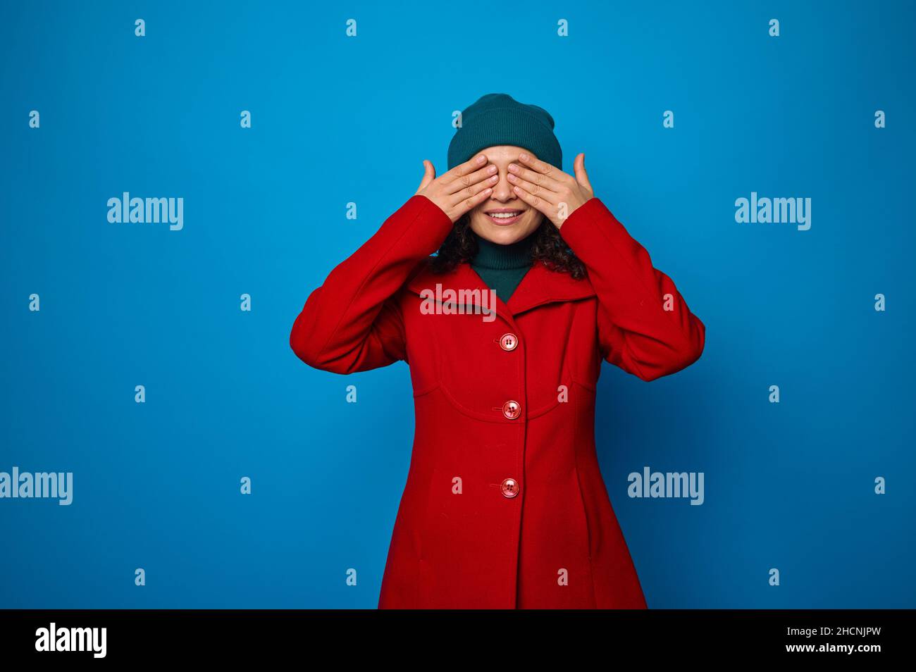 Charming wonderful pretty woman in bright red coat and green hat covering her eyes with hands and smiling with cheerful toothy smile posing against bl Stock Photo