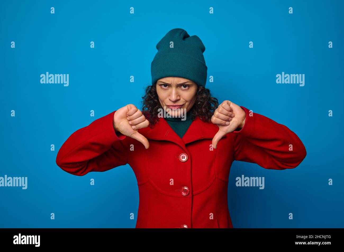 Disappointed upset sad frustrated pretty woman in bright red coat and green hat looking at camera and showing thumbs up, posing against blue backgroun Stock Photo