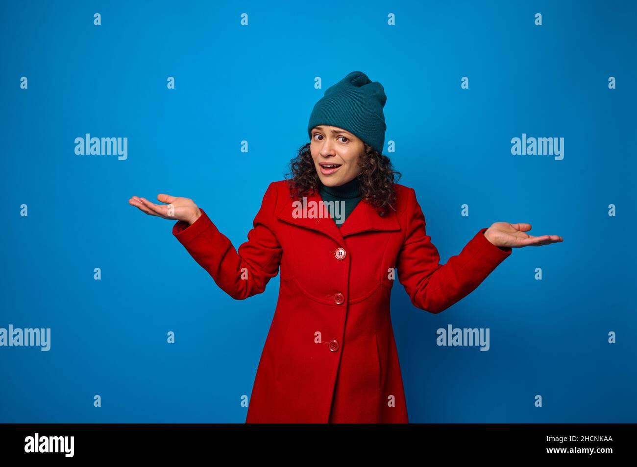 Surprised astonished young curly haired brunette woman in red coat and green hat poses against blue background, looks at camera holding imaginary copy Stock Photo