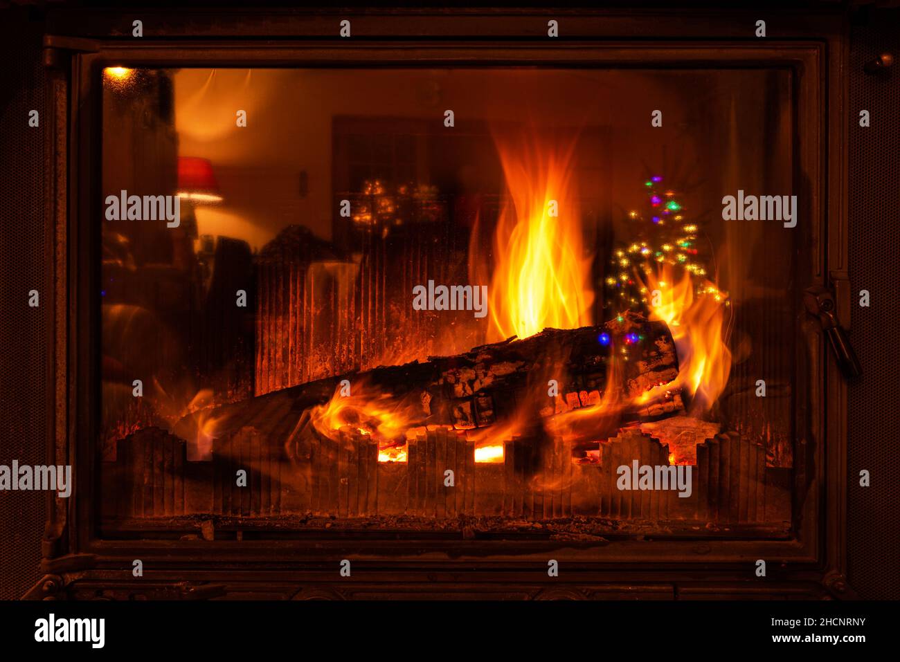 Wood logs burning in a fireplace for Christmas. A Christmas tree decorated with light tinsels is reflected on the glass of the fireplace. Stock Photo