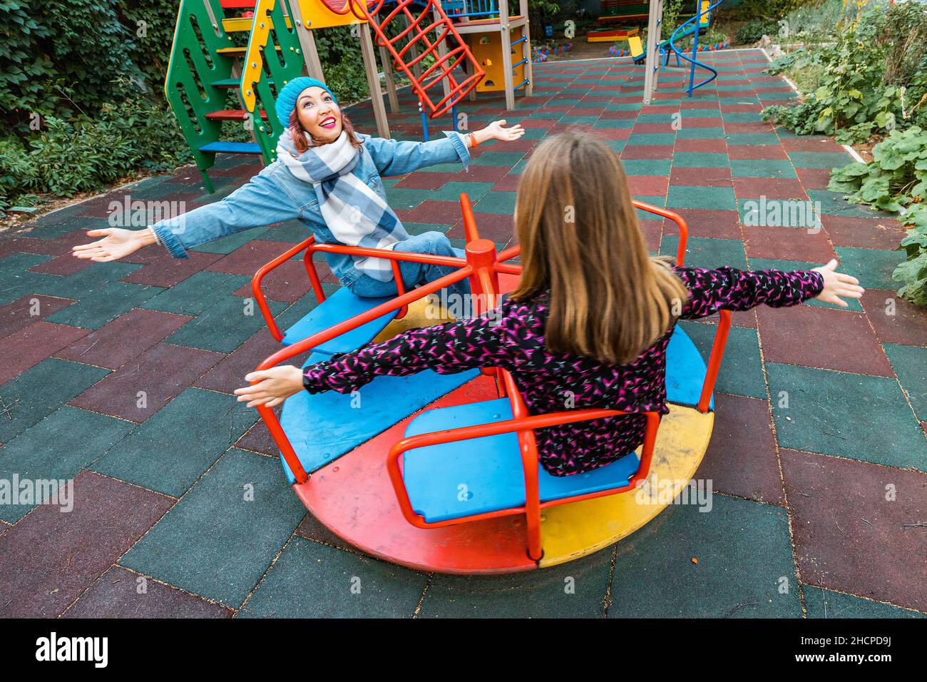 Two childish happy woman friends or teenage girl having fun on seesaw and carousel on a playground. Concept of psychology of the new generation and re Stock Photo