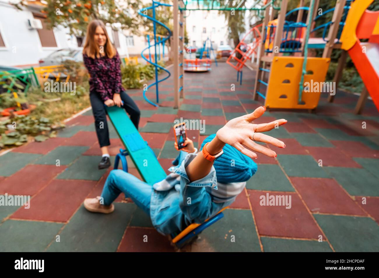 Two childish happy woman friends or teenage girl having fun on seesaw on a playground. Concept of psychology of the new generation and relations Stock Photo