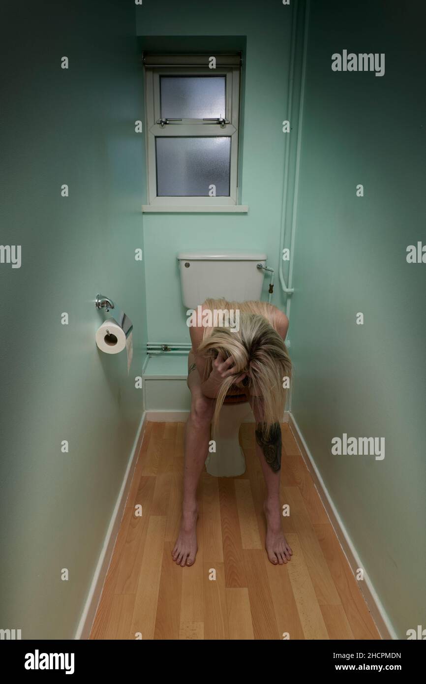 Young woman with long dyed blonde hair and tattooed legs sitting on toilet with head in hands and hair hiding her face as if worried and frustrated Stock Photo