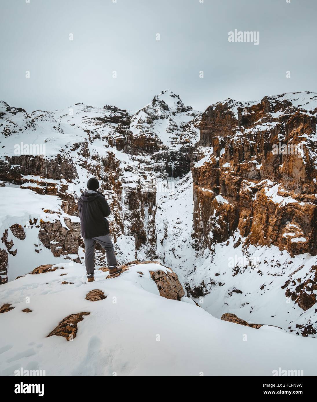 Young man traveler tourist standin in front of Secret canyon named Mulagljufur canyon in South Iceland. Waterfall below mountain top in winter and Stock Photo