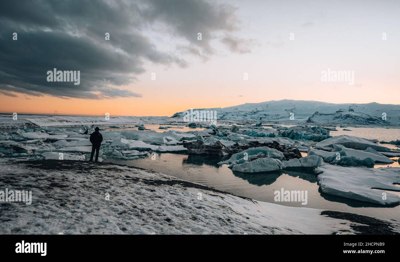 Young tourist standing in front of famous lake Joekulsarlon glacial lagoon and diamond beach with its icebergs and ice floes in Iceland during sunset Stock Photo