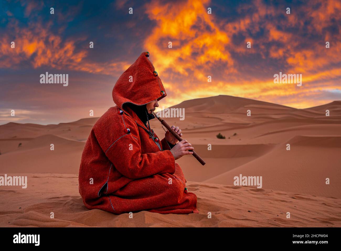 Man in traditional red clothes playing flute while sitting on sand in desert Stock Photo