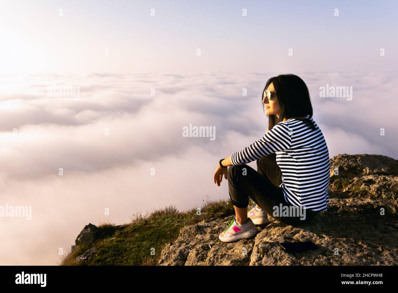 Young caucasian woman sits calmly on the rock and looks left above the clouds. Thoughtfulnessl and solitude concept. Stock Photo