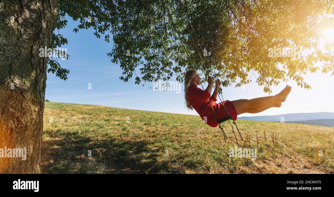 Cheerful lady in red sundress leaning back in her swing in sunny golden countryside. Evening sunshine beaming on lovely girl swaying under single tree Stock Photo
