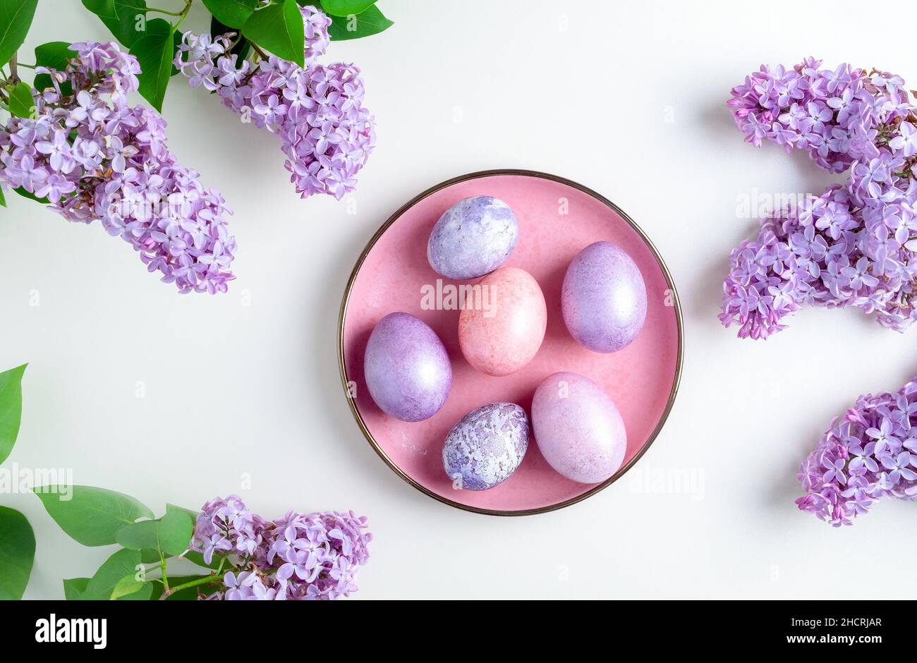 Close-up top view of beautiful nacreous pink and lilac easter eggs lie on a pink plate framed by flowering branches of lilac on a white background. Se Stock Photo