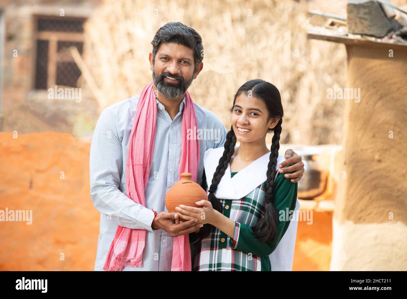 Portrait of happy Indian farmer father and daughter holding clay money box or gullak, Smiling beard man and young girl traditional piggy bank, dad tea Stock Photo