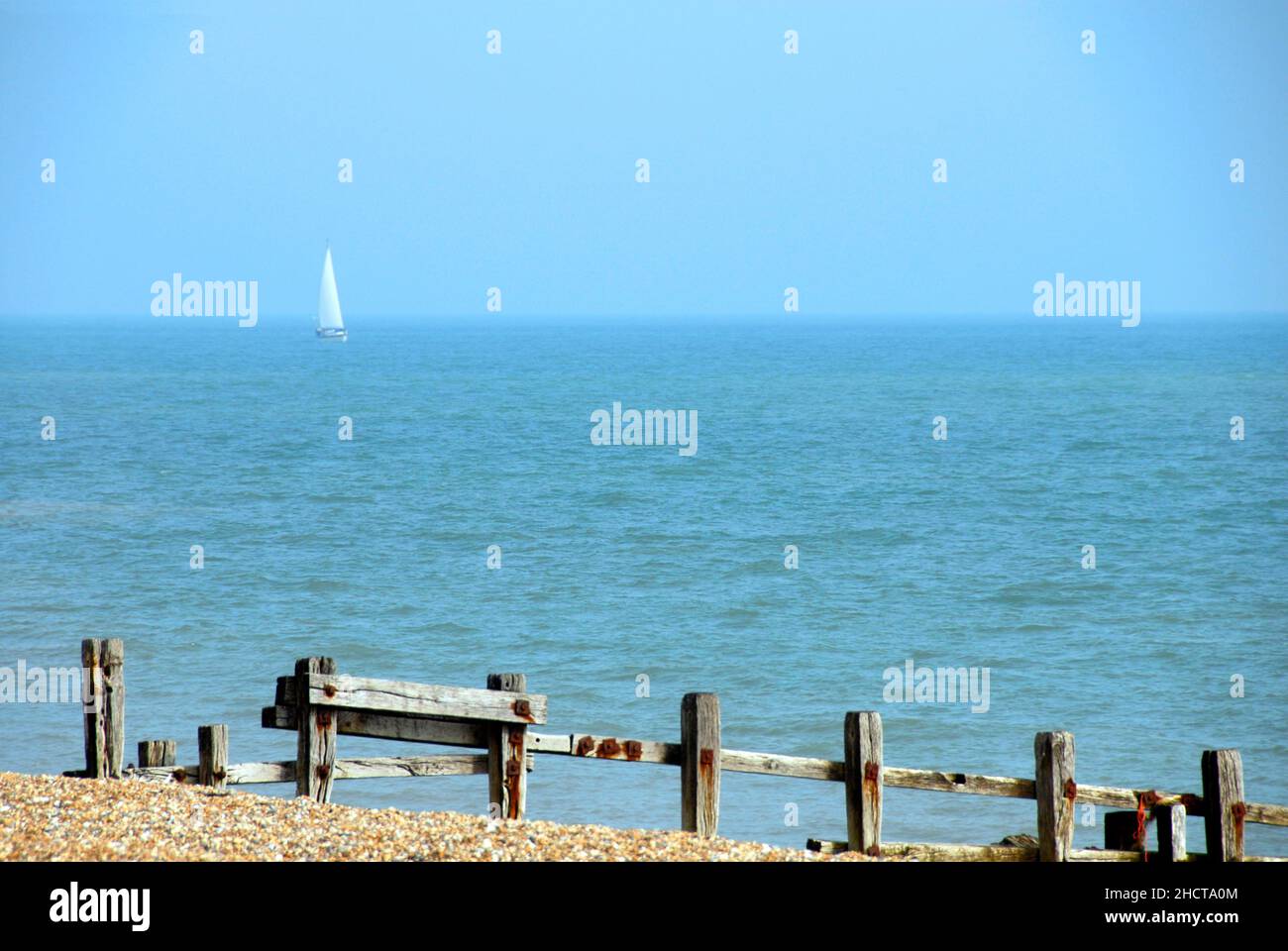 Lone yacht with white sail, Bermuda rig, sailing in the English Channel off Pevensey Bay, East Sussex, England and old wooden breakwater in foreground Stock Photo